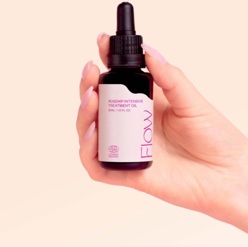 Pieni tumma pullo tipatulla, merkattu 'Rosehip Intensive Treatment Oil' by Flaw. Kevyt ihonsävyinen käsi ja maalatut kynnet.