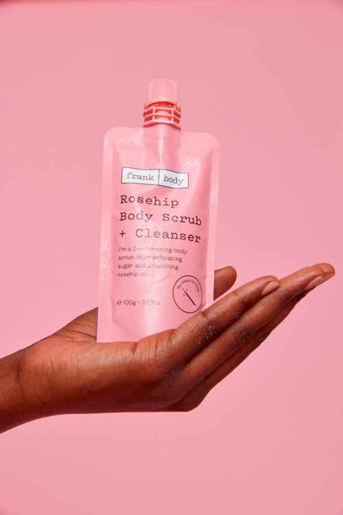 Eine Hand hält eine pinke Tube mit Körperpeeling und Reiniger vor rosa Hintergrund.