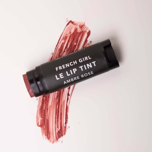 Black lip tint tube on pinkish-red smudge background, labeled 'French Girl Le Lip Tint Ambre Rose'.