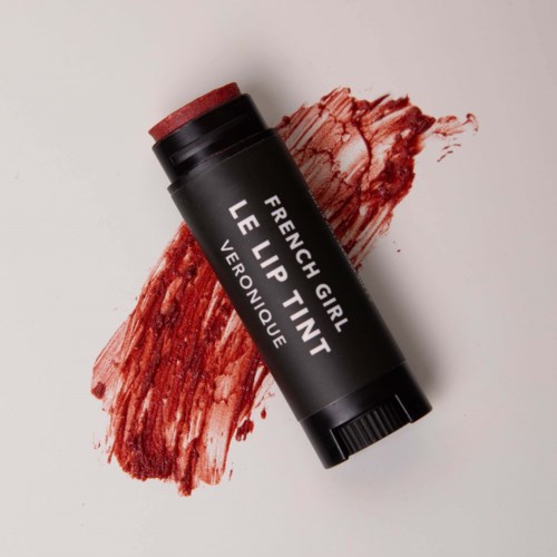 A black tube labeled 'French Girl Lip Tint' lies on a background with red lipstick smudges.