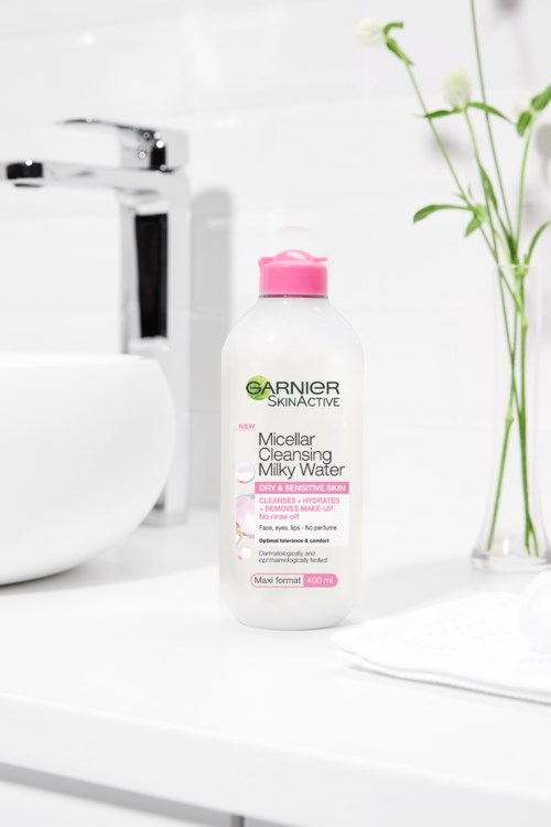 A pink-capped Garnier Micellar Cleansing Milk Water bottle on a white bathroom counter, with a faucet and green plant nearby.