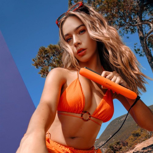 A woman in an orange bikini holds a flat iron, with trees and a mountain in the background on a sunny day.