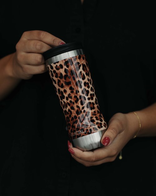 Henkilö pitää leopardimerkkistä termosmukia, jolla on musta paita ja rannerengas, tumman taustan edessä.