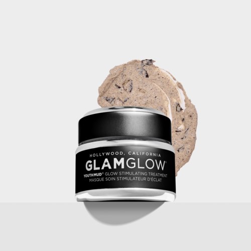 En liten burk med GLAMGLOW Youthmud-maska med ett beige skrubb bakom, på en ljusgrå bakgrund.