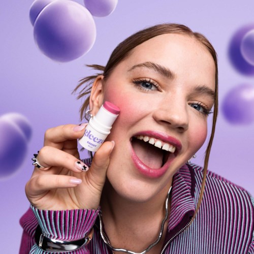 Nainen hymyilee ja pitää pinkkiä ja valkoista huulirasvapakkausta lähellä huuliaan. Violettiä palloja leijuu ympäri.