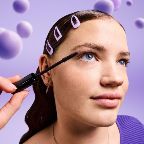Nainen lisää ripsiväriä silmiinsä, taustalla violetit pallot ja violetit hiushokkareet.