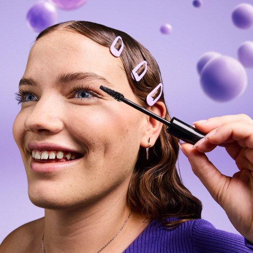 Nainen hymyilee, kun joku laittaa ripsiväriä, hiuspinnit hiuksissa ja violetti tausta palluroineen.