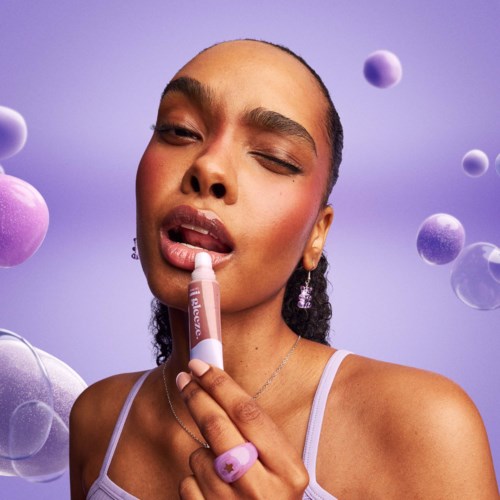 Een vrouw brengt lipgloss aan met half gesloten ogen, omgeven door zwevende paarse en roze bollen tegen een paarse achtergrond.