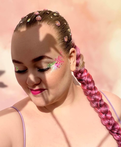 Een vrouw met roze vlecht haar, versierd met gekleurde stenen, heeft glinsterende make-up met bloemen op haar gezicht.