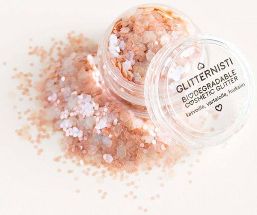 A small clear container of biodegradable glitter with peach and white colors, spilled on a white surface.