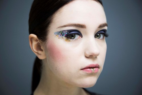 En kvinna med glitter på ögonlocket, rosa rouge och glitter på läpparna, med en neutral bakgrund.