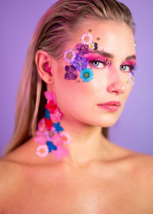 Kvinna med färgglad blom- och glittermakeup i ansiktet och på halsen, mot lila bakgrund.