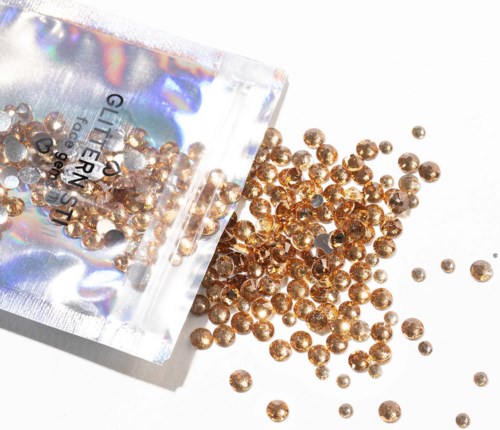 Small round gold-colored beads spilling from a shiny bag on a white surface.