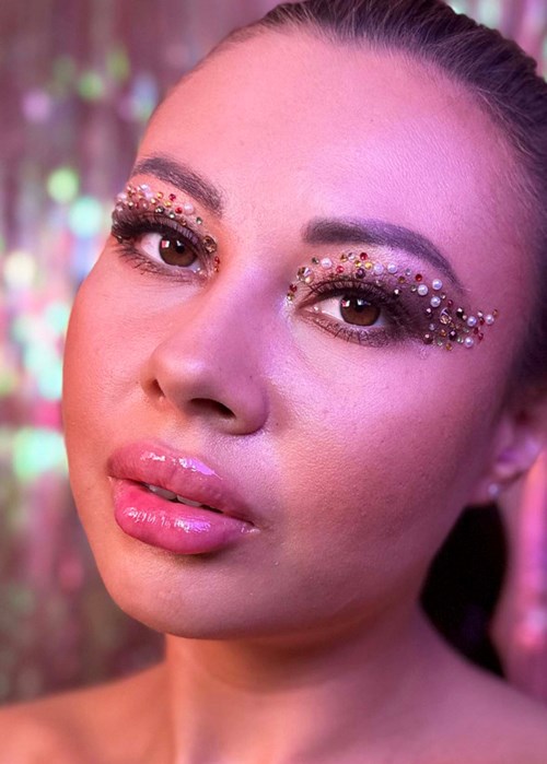 A woman with shiny, gemstone-studded makeup around her eyes and pink glossy lips. Blurred colorful background.