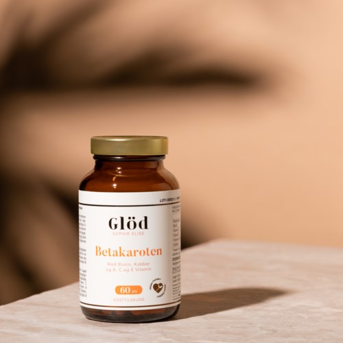 En brun flaska märkt "Betakaroten" med guldkapsel, placerad på en ljus yta. Oskarp ansikte i bakgrunden.