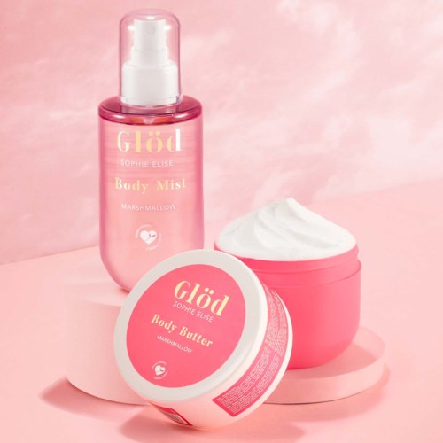 Lyserosa kroppsspray og kroppsmørbeholdere med hvite og rosa etiketter, satt mot en rosa bakgrunn.