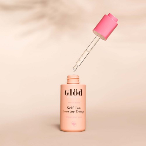 En rosa flaske merket 'Self Tan Booster Drops' med en pipette over den som slipper en dråpe.