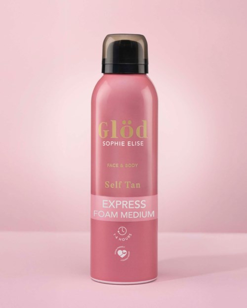Lyserosa sprayflaske med sort topp, merket Glöd Face & Body Self Tan Foam Medium, på rosa bakgrunn.