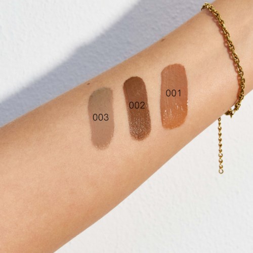 Swatch av tre olika foundationnyanser på en persons arm, numrerade 001, 002 och 003, bredvid ett guldarmband.