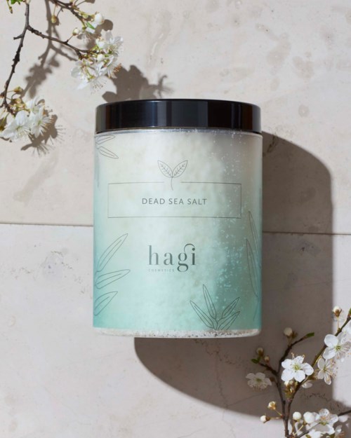 En beholder mærket 'Dead Sea Salt' på en lys overflade, omgivet af hvide blomster med skyggeeffekter.