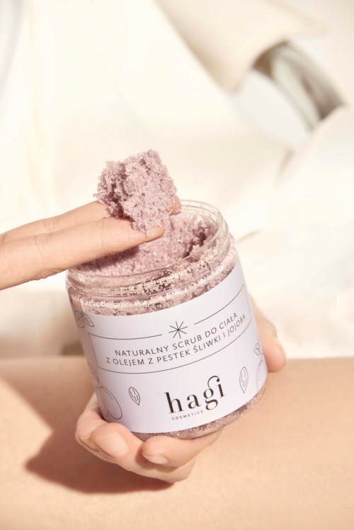 A hand holds a jar of purple body scrub with a scoop of the scrub. The jar has a white label with text.