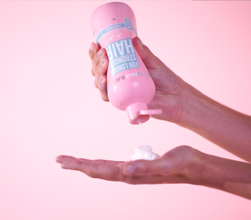 Hand som håller en rosa flaska, trycker ut skum på handflatan mot en rosa bakgrund.