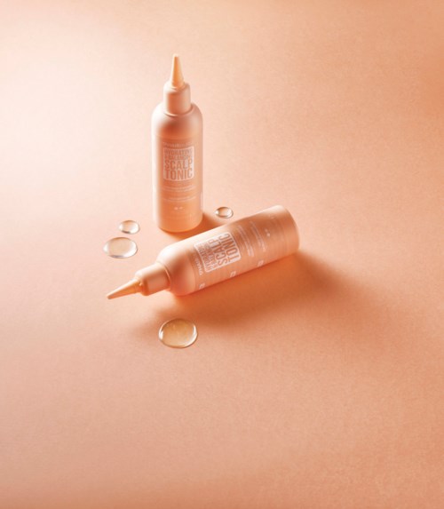 Twee kleine perzikkleurige flesjes met druppelaars op een beige oppervlak, met wat heldere vloeistofdruppels eromheen.