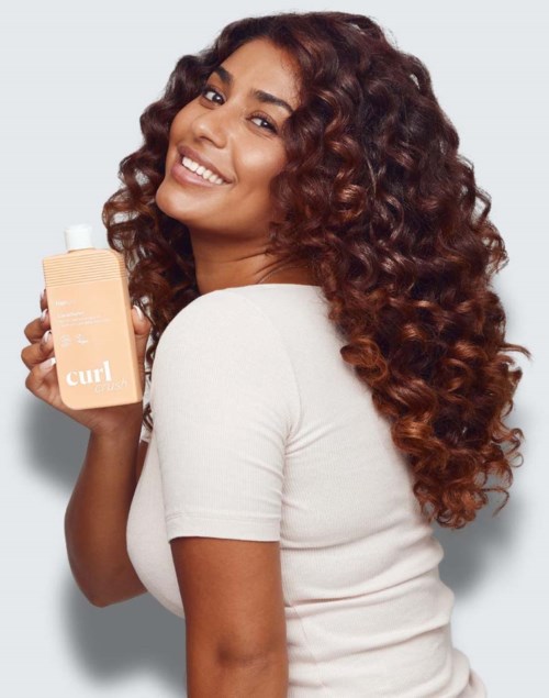 Vrouw met krullend haar die een fles Curl-shampoo vasthoudt en glimlacht naar de camera.