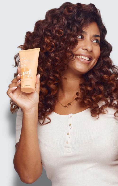 Glunderende vrouw met krullend haar die een perzikkleurige fles haarverzorgingsproduct vasthoudt.