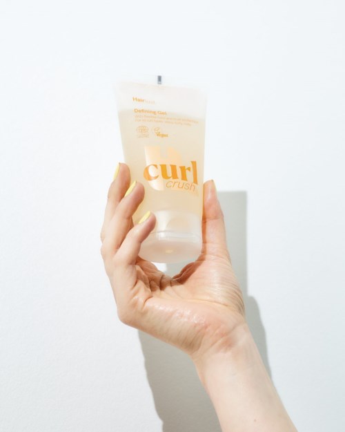 Hånd som holder en klar tube med curl-definerende krem og oransje tekst.
