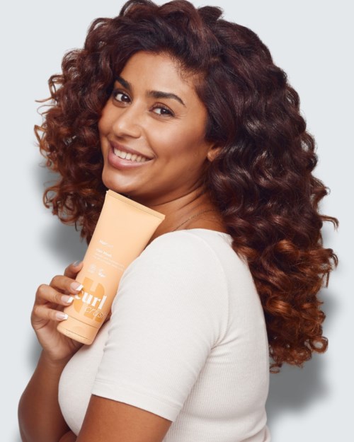 Lachende vrouw met krullend haar die een beige huidverzorgingsbuis vasthoudt, wit shirt.