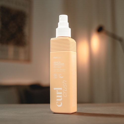 Beige hårpleiesprayflaske med sprayhode på en treoverflate, uskarp varm bakgrunn.