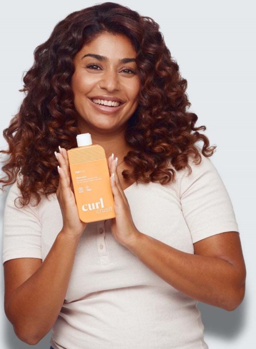 Vrolijke vrouw die een fles curl cream haarproduct vasthoudt, draagt een wit shirt, heeft krullend haar en een grijze achtergrond.