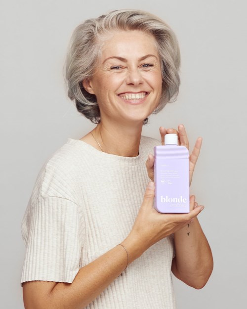 lachende vrouw met grijs haar die een lavendelkleurige fles vasthoudt met Blonde erop.