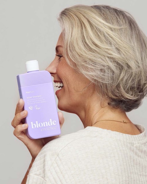Lachende oudere vrouw met grijs haar die een paarse shampoo flesje met blond label vasthoudt.