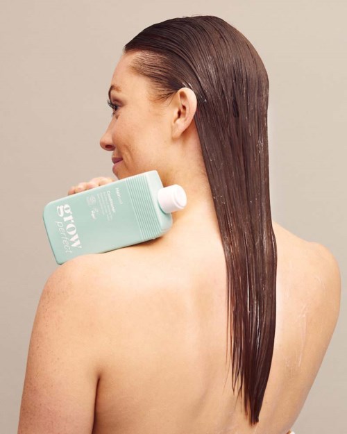Vrouw met lang, steil bruin haar die een lichtblauwe shampoo fles op haar schouder houdt.