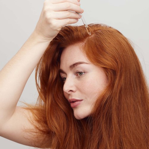 Een vrouw met lang, rood haar gebruikt een pipet om serum op haar hoofdhuid aan te brengen. Ze kijkt naar beneden en heeft een neutrale uitdrukking.