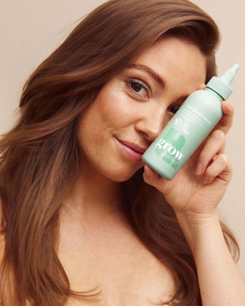 Een vrouw met lang bruin haar houdt een lichtgroene sprayfles dicht bij haar gezicht en glimlacht zacht.