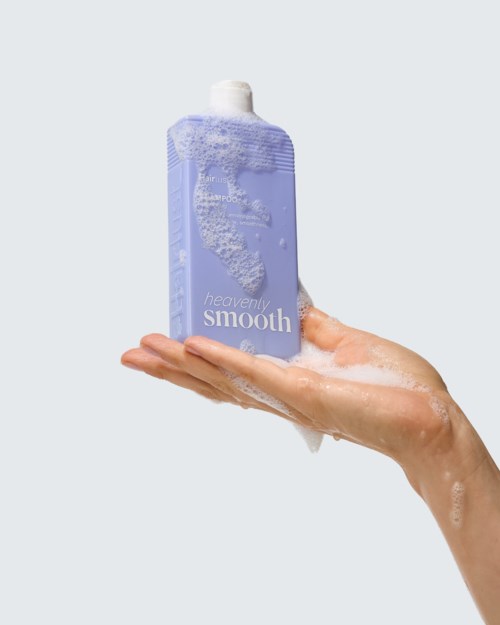 Een hand houdt een lavendelkleurige douchegelverpakking met schuim op het oppervlak en een milde zeepvlag. Het label zegt heavenly smooth.
