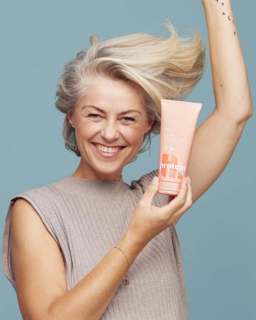 Smiling woman with gray hair holding a pink skincare product tube, her hair lifted and a blue background.