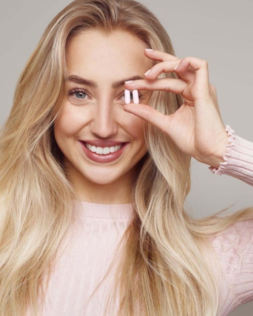 Een lachende vrouw met lang blond haar houdt twee roze pillen dichtbij haar oog.