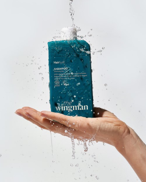 A hand holding a bottle of teal shampoo with water splashing around it.