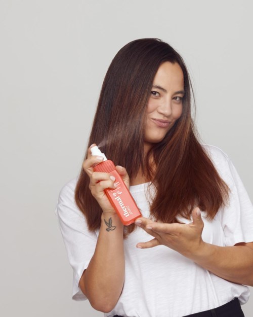 A woman with long brown hair holds a spray bottle labeled hair product, smiling at the camera.