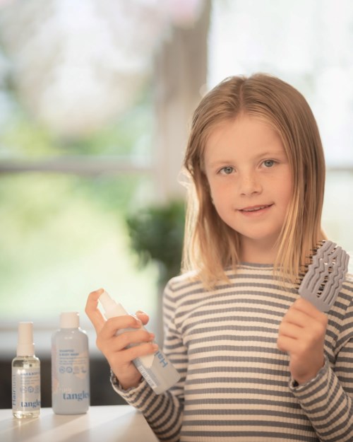 Jong meisje met blond haar dat een haarborstel en een sprayflesje vasthoudt, naast haarverzorgingsproducten.