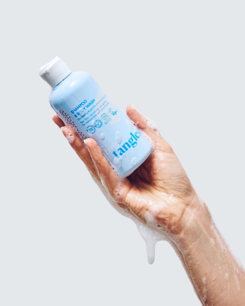 Hand vasthoudend een blauwe shampoo fles met schuim op hand, tegen een eenvoudige grijze achtergrond.