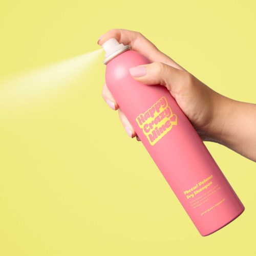 En hand sprayar rosa hårspray med en gul etikett som säger "Happy Crazy Time" i gult text på en gul bakgrund.