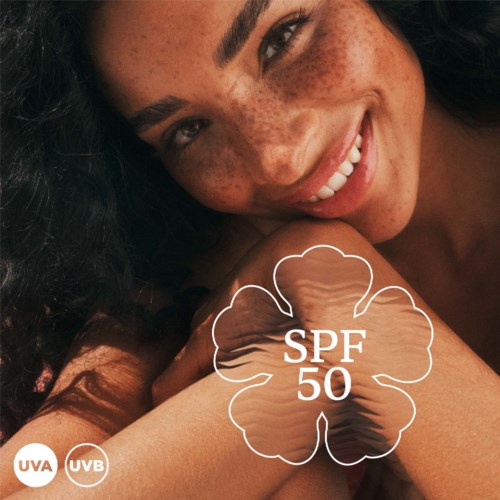 En leende kvinna med fräknar visar sitt ansikte och arm med SPF 50 solskydd. Bakgrunden är mörk.