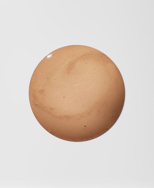 En rund, brun makeup-svamp med nogle darker pletter, placeret på en hvid baggrund.