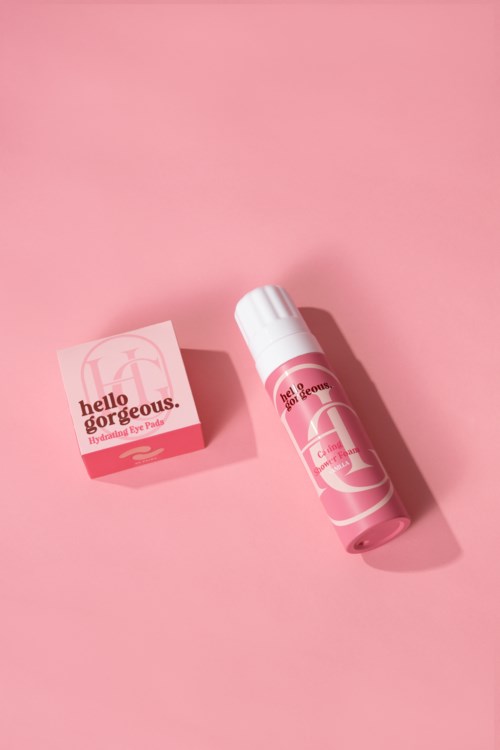 Ljusrosa hudvårdsprodukt med tub och box, båda märkta "Hello Gorgeous." Bakgrunden är rosa.