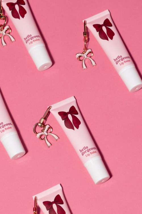 Pink lip balm tubes with red bows and small pink bow charms, arranged diagonally on pink background.